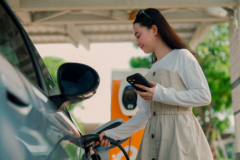 Woman stands beside an electric vehicle at an outdoor charging station, plugging in the charging cable while checking her smartphone. The image shows EV charging infrastructure in use, highlighting smartphone-enabled charging, clean energy, and everyday adoption of electric vehicles.