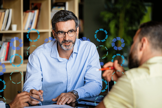 A man wearing glasses and a blue shirt talking to a colleague while sat at a table